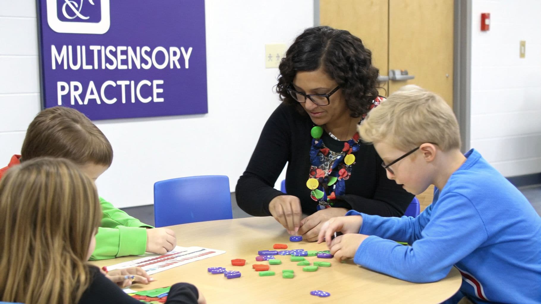An adult leads three young children in a hands-on multisensory learning activity with colorful pieces.