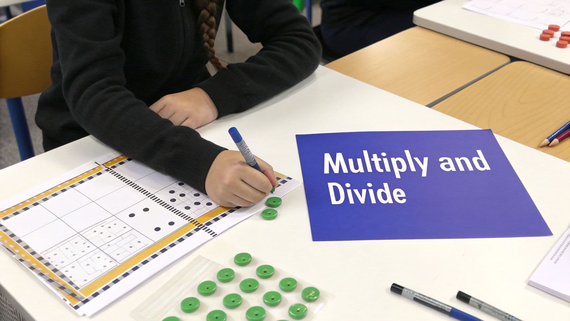 A student's hands work on a math worksheet with green counters and a 'Multiply and Divide' sign.