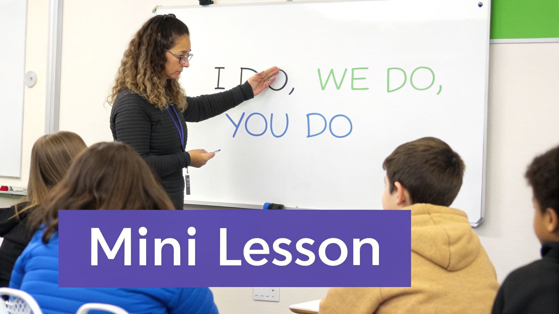 A teacher points to a whiteboard displaying 'I DO, WE DO, YOU DO' during a classroom lesson.
