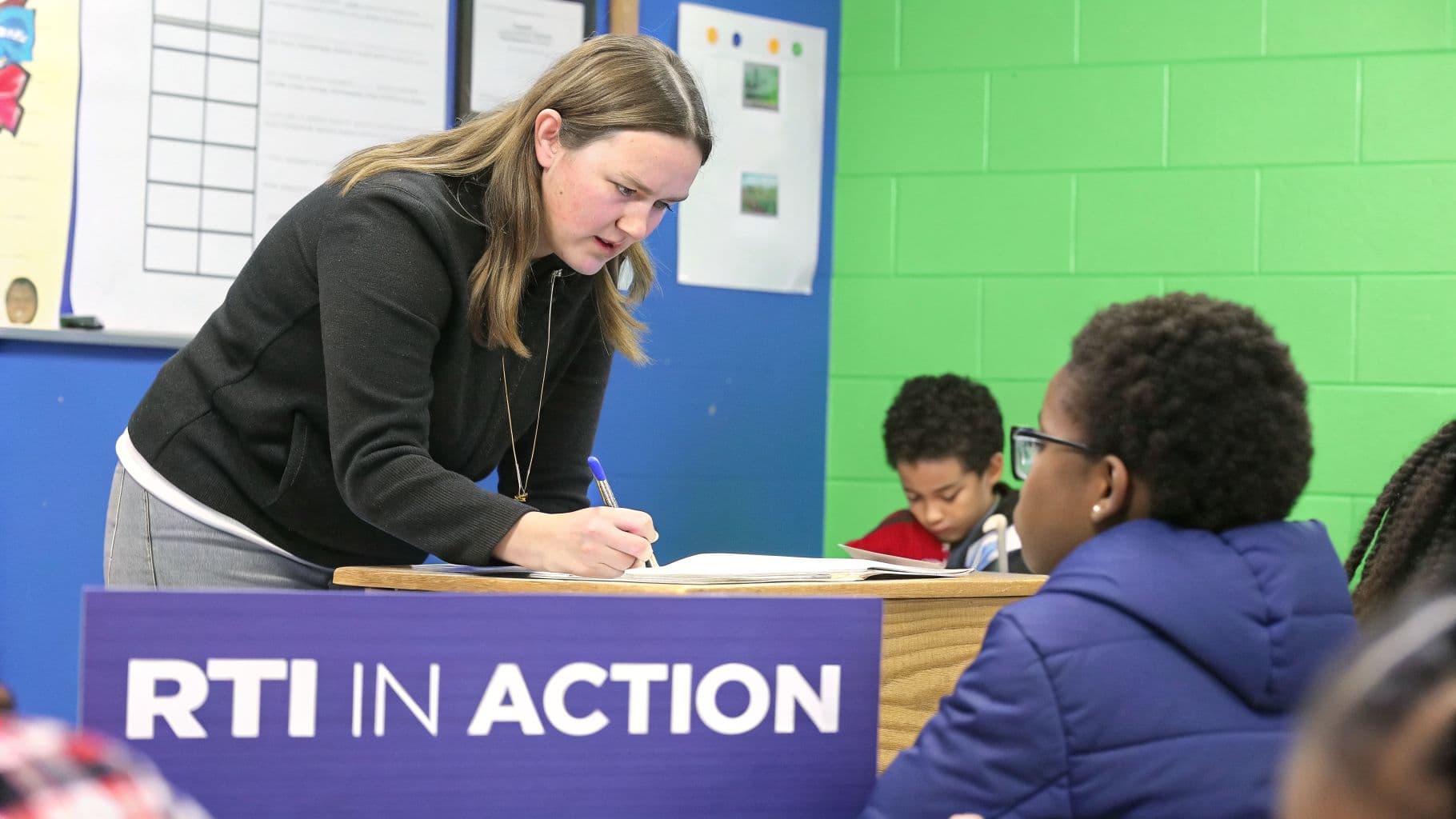 An educator writes in a notebook, assisting students in a classroom with a 'RTI in Action' sign.