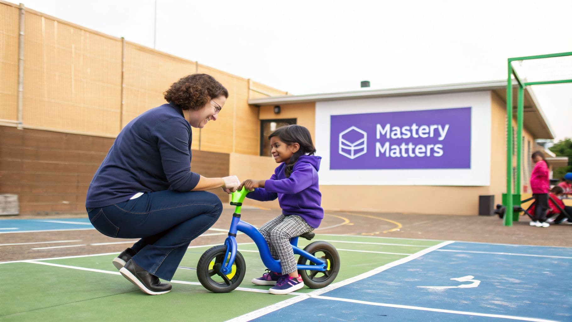 A teacher helps a young student on a blue balance bike, promoting mastery in learning.