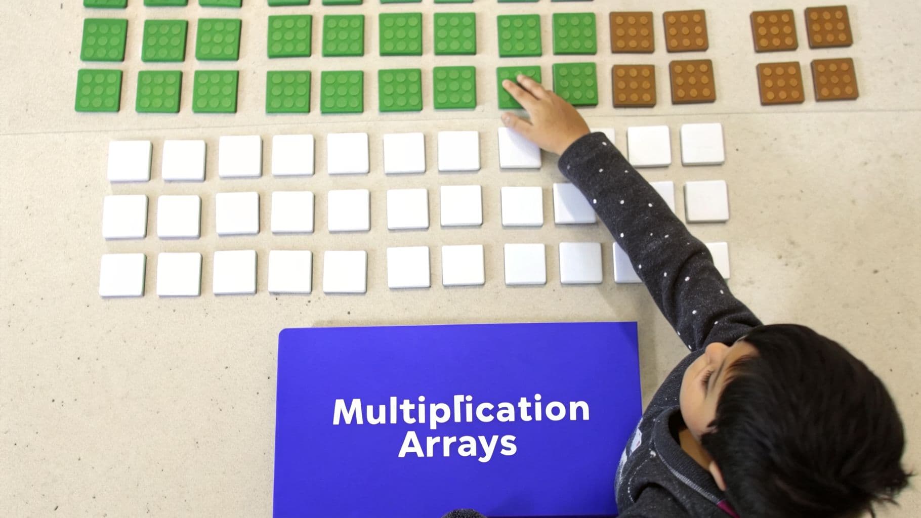 A child's hand places a white tile next to green and brown blocks, illustrating multiplication arrays.